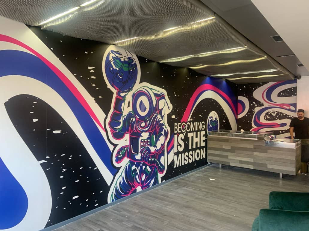 a man standing behind a desk and an artistic wall mural of an astronaut  with the world in his hands floating on the space a man standing behind a desk and an artistic wall mural of an astronaut with the world in his hands floating on the space