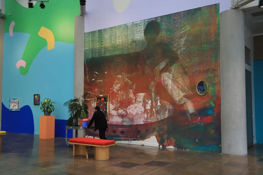 A  gallery or museum with a huge custom wall mural featuring  an aquarela painting with a man sitting in one side of a sofa and a low round table with several items in the middle A gallery or museum with a huge custom wall mural featuring an aquarela painting with a man sitting in one side of a sofa and a low round table with several items in the middle