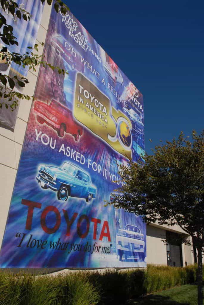 A vividly colorful billboard for toyota, featuring various car models and slogans like "oh what a feeling!" and "i love what you do for me," set against a bright blue sky, next to a building with trees in the foreground. A vividly colorful billboard for toyota, featuring various car models and slogans like "oh what a feeling!" and "i love what you do for me," set against a bright blue sky, next to a building with trees in the foreground.
