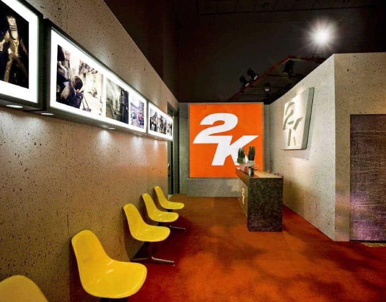 An art gallery interior with vibrant orange walls featuring a large "2k" logo. grey concrete partitions separate sections, decorated with framed black-and-white photographs. yellow modern chairs line the space, illuminated by spotlights. An art gallery interior with vibrant orange walls featuring a large "2k" logo. grey concrete partitions separate sections, decorated with framed black-and-white photographs. yellow modern chairs line the space, illuminated by spotlights.