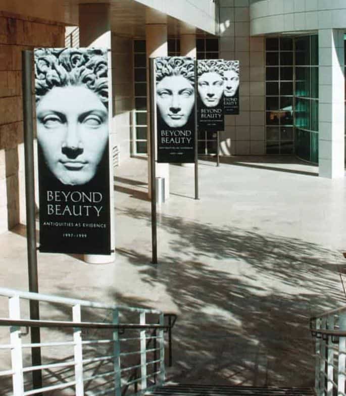 Three promotional banners for an art exhibit titled "beyond beauty: antiquities as evidence" stand in front of a modern building with glass doors and dappled sunlight on the floor. Three promotional banners for an art exhibit titled "beyond beauty: antiquities as evidence" stand in front of a modern building with glass doors and dappled sunlight on the floor.
