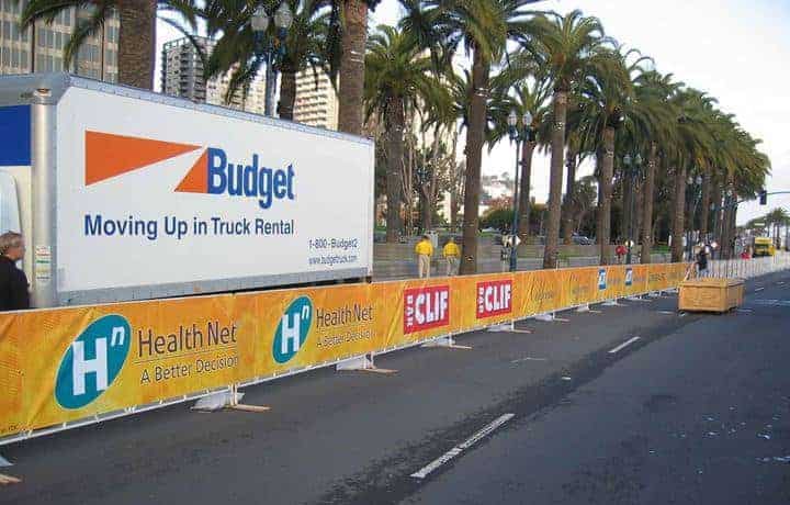 A city street lined with palm trees and barrier fences displaying various sponsor banners including "budget", "health net", and "clif bar". the road appears to be prepared for an upcoming event, possibly a race or parade. A city street lined with palm trees and barrier fences displaying various sponsor banners including "budget", "health net", and "clif bar". the road appears to be prepared for an upcoming event, possibly a race or parade.