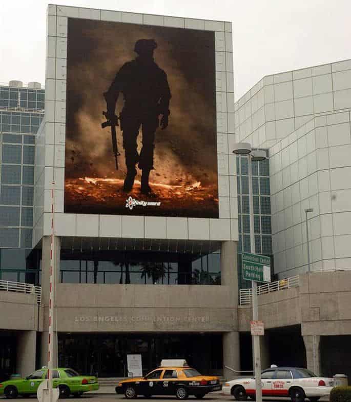 A billboard depicting a silhouette of a soldier standing in a smoky battlefield is displayed on the exterior of the los angeles convention center, flanked by modern buildings and colorful parked cars. A billboard depicting a silhouette of a soldier standing in a smoky battlefield is displayed on the exterior of the los angeles convention center, flanked by modern buildings and colorful parked cars.