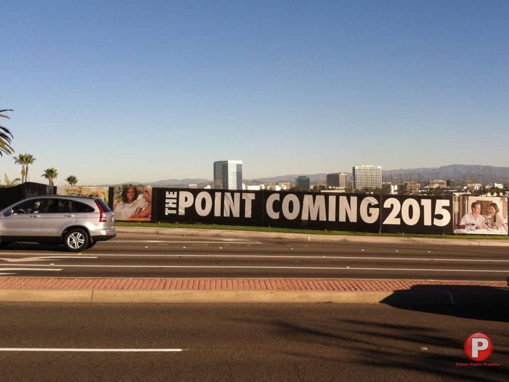 A large construction banner along a wide, multi-lane road with palm trees and a car passing by displays the text 