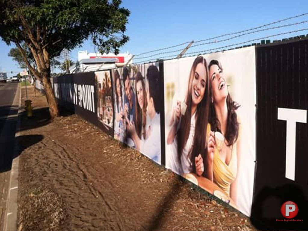 A long black fence with a large banner with happy group photos displayed side by side depicting people laughing and eating. The fence is beside a dirt path and street, with trees and a barbed-wire top.>
            <img decoding=
