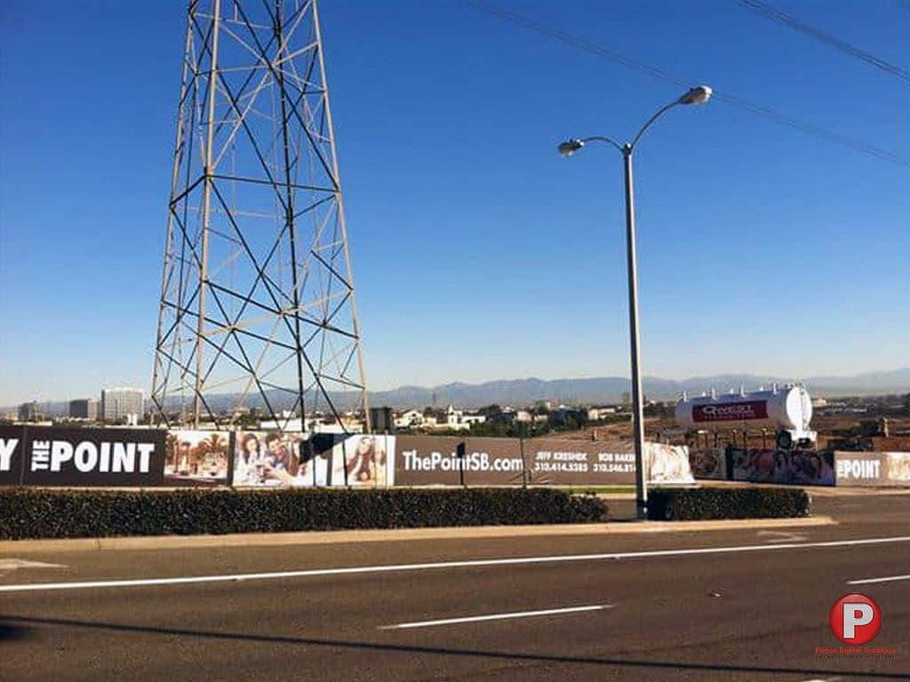 A street view featuring a large fence banner advertisement for 'The Point' with a URL, 'ThePointSB.com,' and contact numbers. The construction site is positioned near a power line tower and a streetlight, with mountains visible in the background under a clear blue sky.
