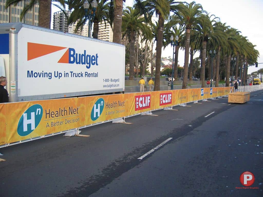 A street barricaded by a temporary fence with bright yellow banners displaying Health Net logos. A white Budget truck is parked alongside the barriers, with palm trees and tall buildings in the background.