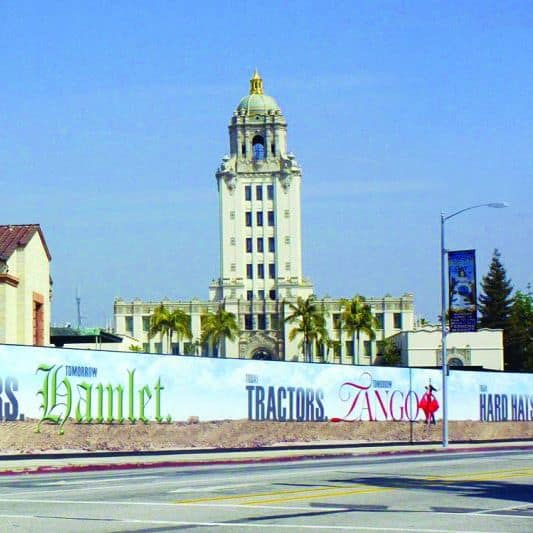A tall, beige clock tower with a dome and golden accents stands in the background, surrounded by palm trees. In the foreground, a long white fence features large Platon Graphics advertisements for "Hamlet" and "Tango." A street with a crosswalk is visible in front of the fence.