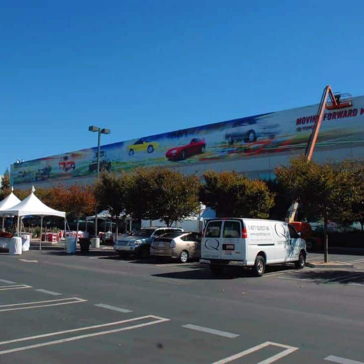 A large construction banner by Platon Graphics featuring speeding cars is displayed on the side of a building against a clear blue sky. Below it, a white van and several cars are parked near small trees and white tents, indicating an event setup in the parking lot of Toyota. A boom lift is positioned near the mural.
