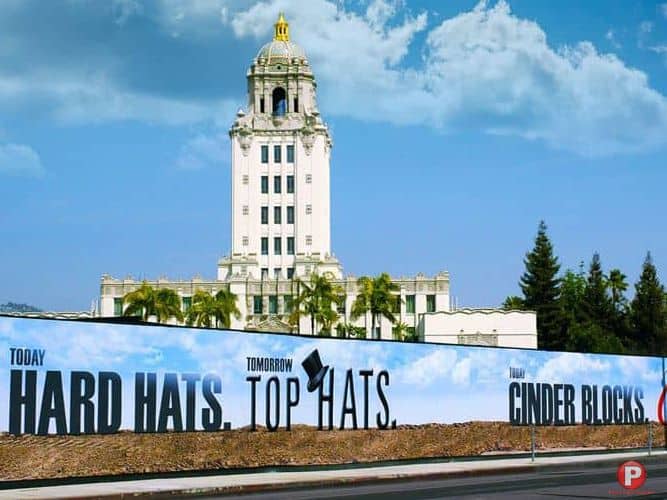 A tall white building with a clock tower stands under a blue sky with clouds. In front, a large construction fence banner displays text that reads: "Today Hard Hats," "Tomorrow Top Hats," and "Build Cinder Blocks." The construction area is surrounded by dirt and construction materials. Trees and palm trees are visible behind the fence.